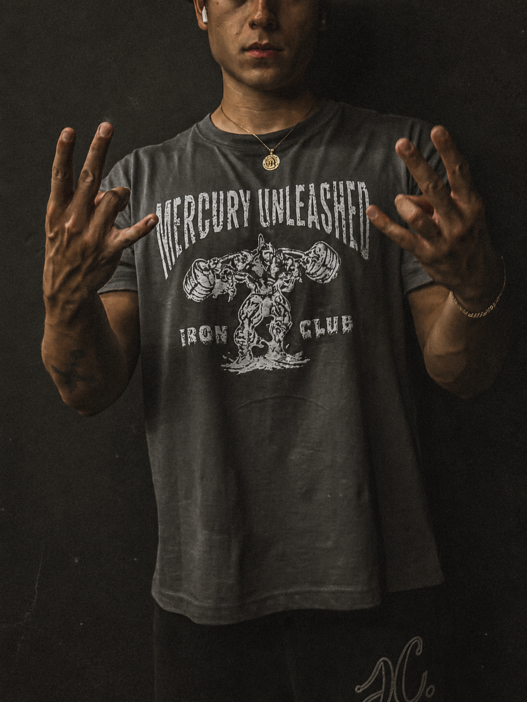 A man wearing a dark gray graphic t-shirt with a distressed white print that reads 'MERCURY UNLEASHED' and 'IRON CLUB' with a muscular figure graphic. He is posing with his hands raised against a dark, textured background.
