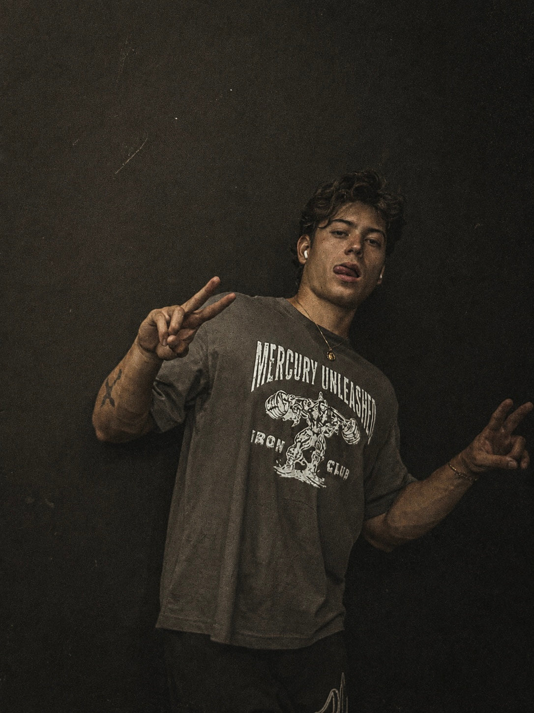 A man posing against a dark textured wall wearing a dark gray t-shirt with a white graphic print that reads 'MERCURY UNLEASHED IRON CLUB'.