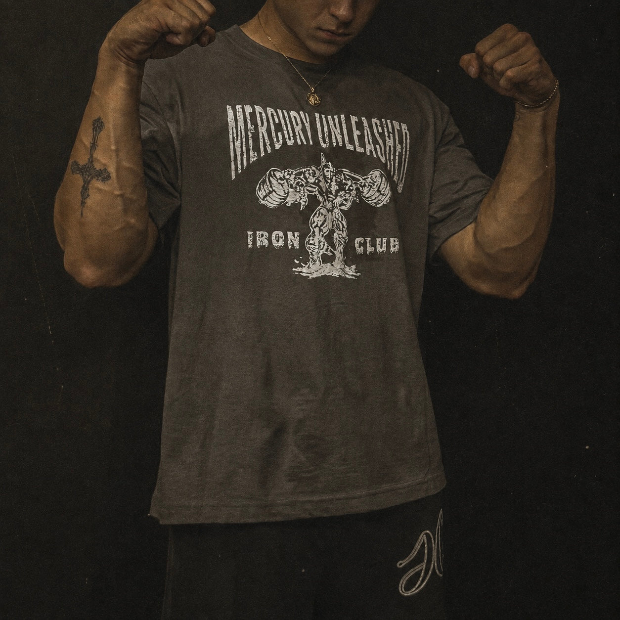 A man wearing a dark charcoal gray t-shirt with a distressed white graphic that reads 'MERCURY UNLEASHED IRON CLUB' and features an illustration of a muscular figure. He is posing with flexed biceps against a dark background.